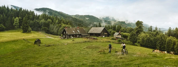 Zwei Radfahrer fahren über eine grüne Wiese in Richtung eines traditionellen Bauernhofs inmitten einer bergigen, bewaldeten Landschaft. Im Hintergrund sind bewaldete Hügel mit Nebelschwaden und mehrere Holzhäuser zu sehen. Rechts grasen Kühe auf einer Weide. Der Himmel ist leicht bewölkt mit blauen Abschnitten.