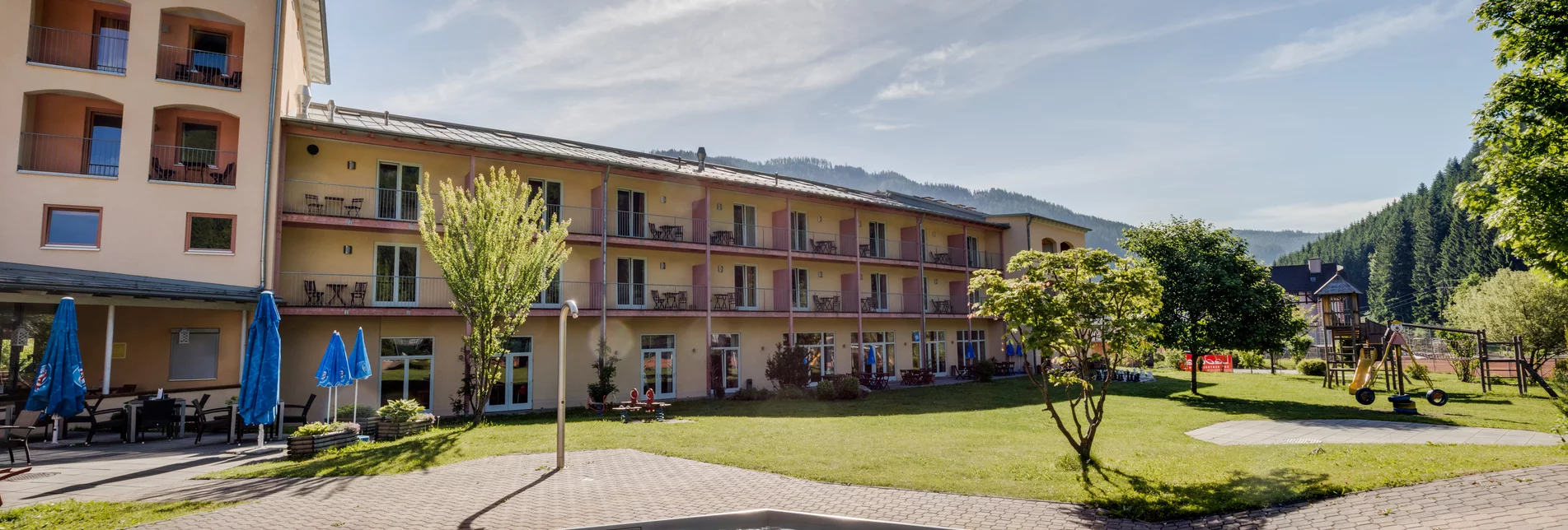 Hotelansicht im Sommer mit großer Wiese, mehreren Bäumen, Kinderbecken und Kinderspielplatz vor dem Haus