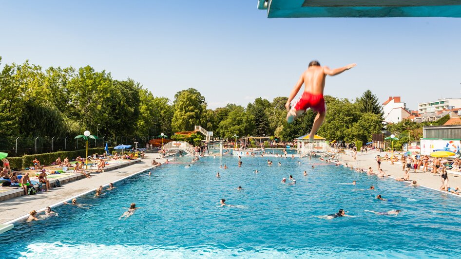 Ein Junge springt ins Schwimmbecken des Augartenbads in Graz an einem sonnigen Tag.