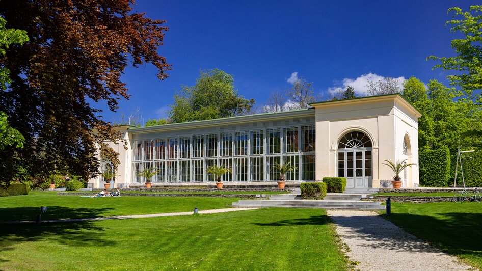 Ein Glashaus im Burggarten in Graz, umgeben von Grün und blauem Himmel.