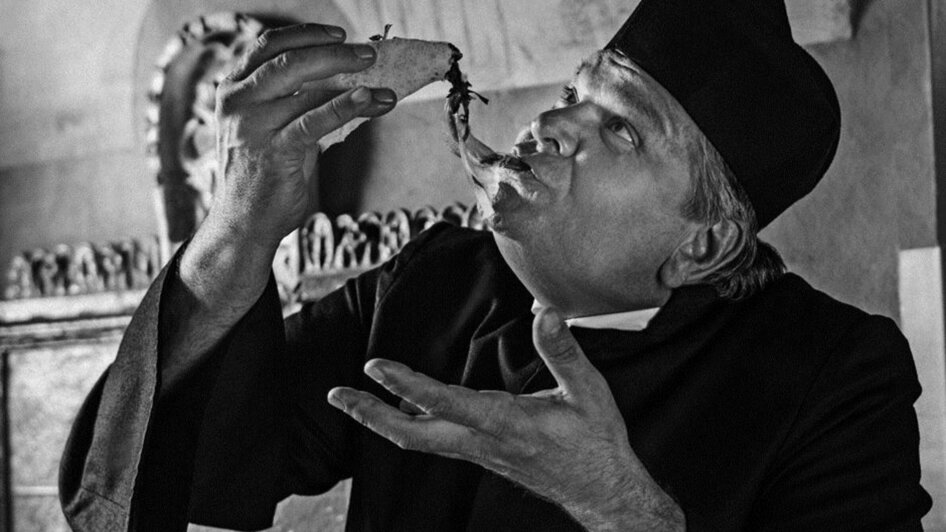 Ein Mann im schwarzen Gewand genießt ein Essen. Das Foto ist schwarz-weiß. | © Don Camillo