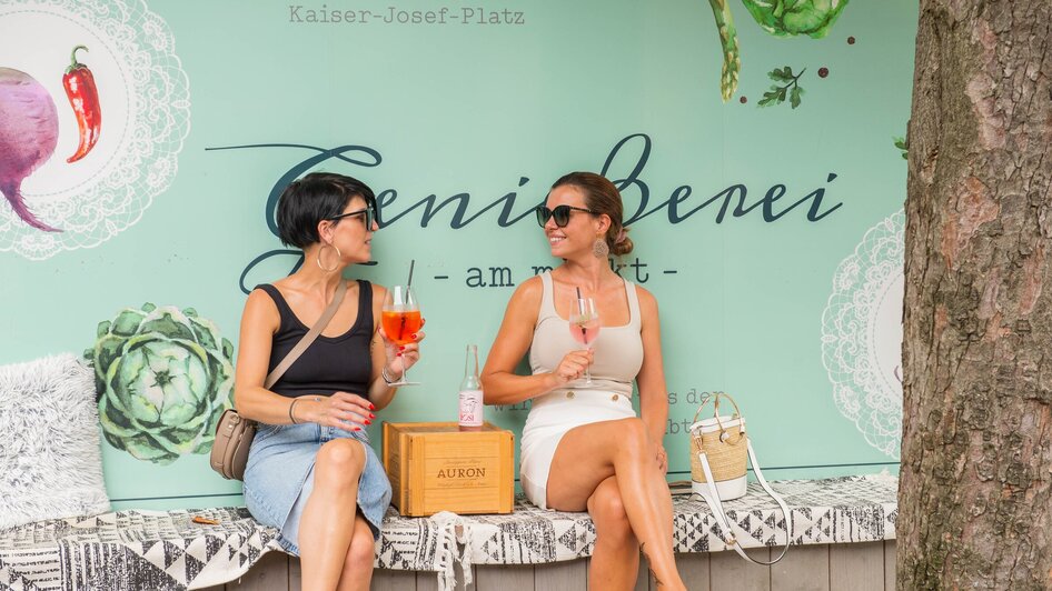 Genießerei am Markt in Graz | © 5komma5sinne - Helmut Schweighofer Zwei Frauen genießen Drinks in Graz vor einer Wand mit Schriftzug. | © 5komma5sinne - Helmut Schweighofer