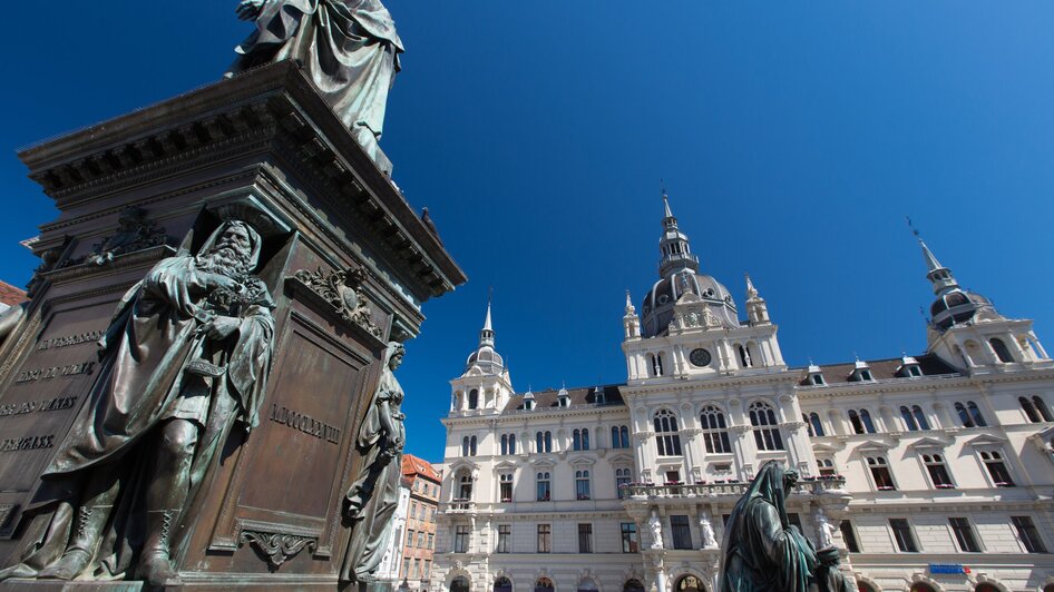 Blick auf das Rathaus am Hauptplatz in Graz mit Statuen im Vordergrund.