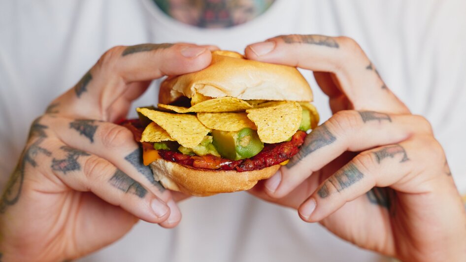 Ein Mann hält einen Hamburger mit Nachos und Avocado-Stücken in seinen Händen. | © Samuel Kuro