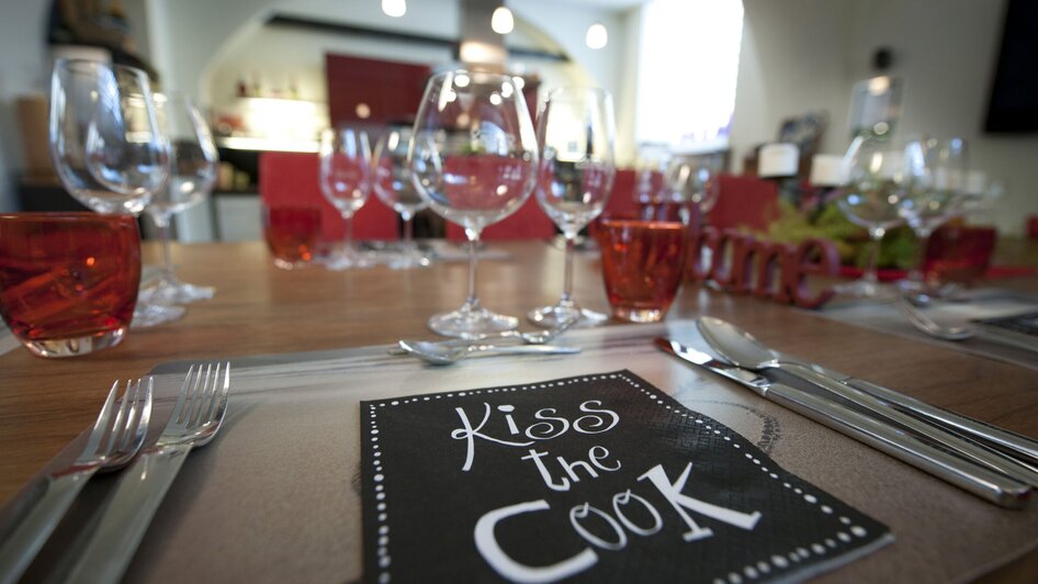 Ein schön gedeckter Tisch mit Gläsern und Tellern und einer Serviette mit der Aufschrift 'Kiss the Cook'. | © kitchen 12 - Christian Jungwirth