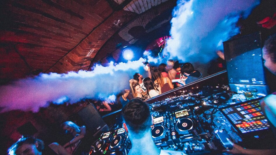 Ein DJ spielt in einem Club, umgeben von Nebel und feiernden Gästen.