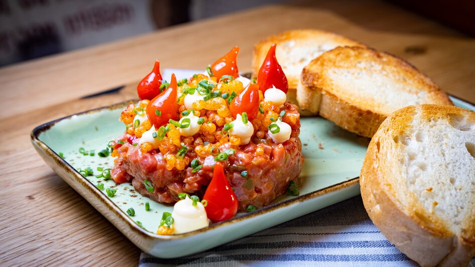 Ein Teller mit Beef Tatar, dekoriert mit Tomaten und Schnittlauch, dazu Weißbrot. | © Kreuzwirt am Rosenberg in Graz