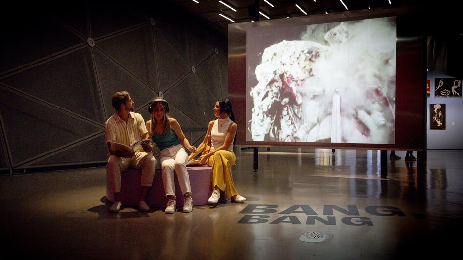 Drei Personen im Kunsthaus Graz, vor einem Video mit dem Text BANG. BANG.