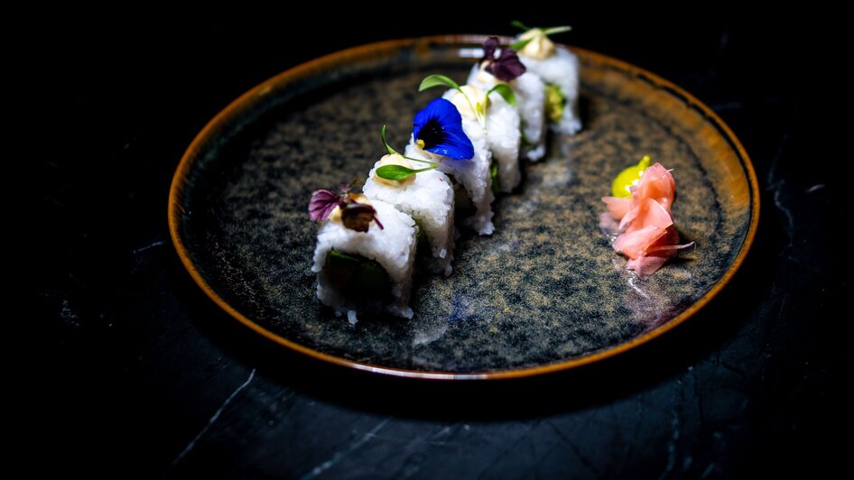 Kunstvolles Sushi mit essbaren Blumen und Ingwer auf schwarzem Teller.
