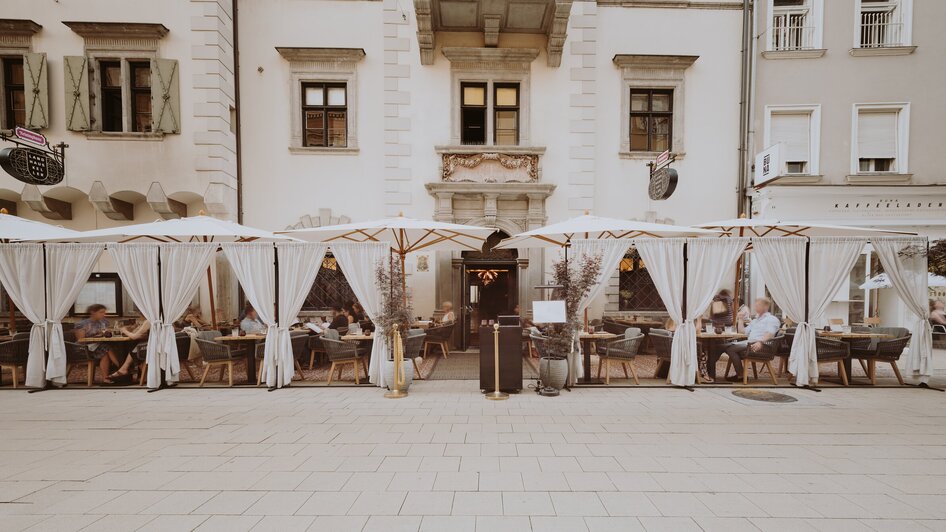 Außenansicht eines Restaurants in Graz mit Sitzgelegenheiten unter weißen Sonnenschirmen.