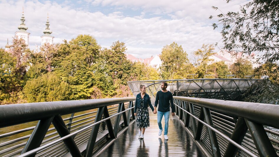 Paar spaziert auf der Murinsel in Graz, umgeben von Bäumen. | © Graz Tourismus