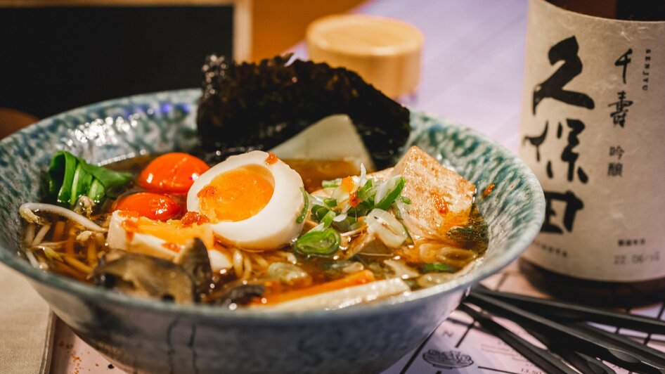 Eine Schüssel Ramen mit Ei, Tofu und Gemüse sowie einer Flasche Sake. | © Moriwano Group - jack-coleman.com
