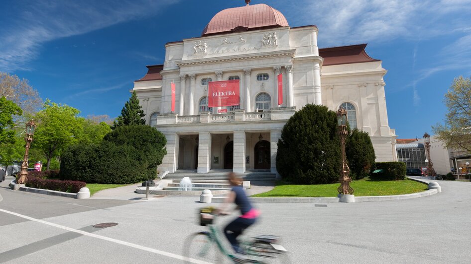 Die Oper Graz mit lebendigem Umfeld und einem Radfahrer auf der Straße.