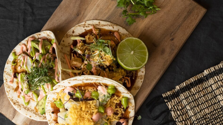 Drei Tacos auf einem Holzbrett mit Limette und frischen Kräutern. | © Jauk Katharina