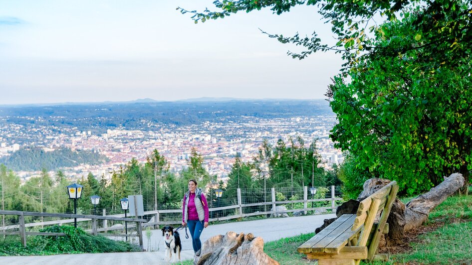 Eine Frau geht mit ihrem Hund auf einem Weg mit Aussicht über Graz.