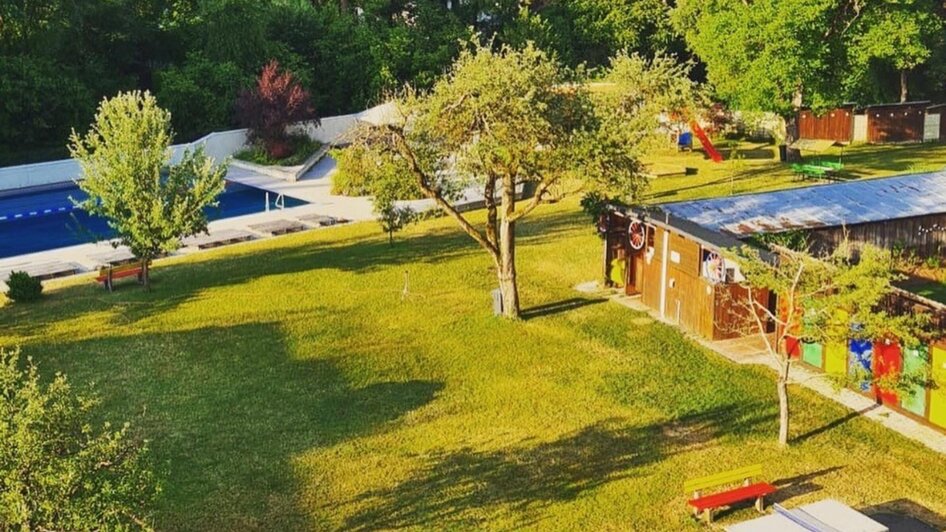 Grüne Wiese mit Picknicktischen und Schwimmbad im Hintergrund.