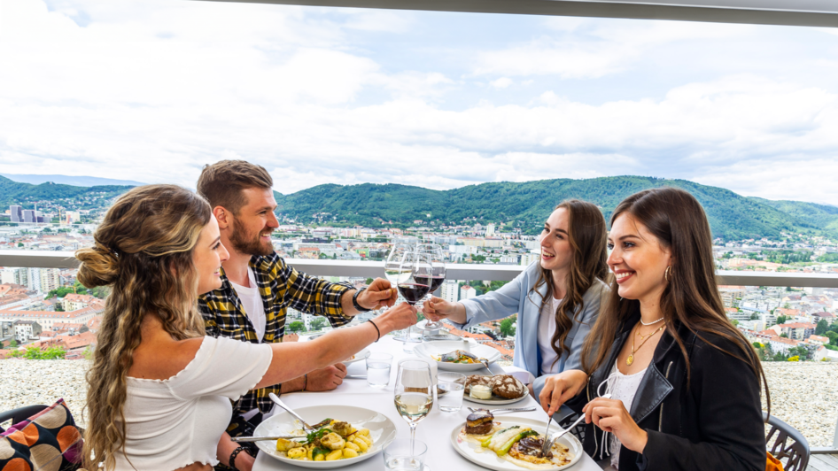 Im Schlossberg Restaurant sitzen vier Personen um einen Tisch mit Blick auf Graz. Sie stoßen mit Weingläsern an und genießen ein stilvolles Essen in einer modernen Umgebung.