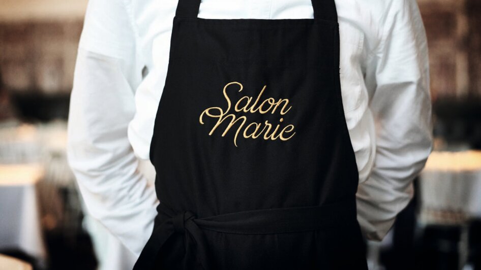 Ein Mann in einem weißen Hemd trägt eine schwarze Schürze mit dem Text 'Salon Marie'. | © Salon Marie
