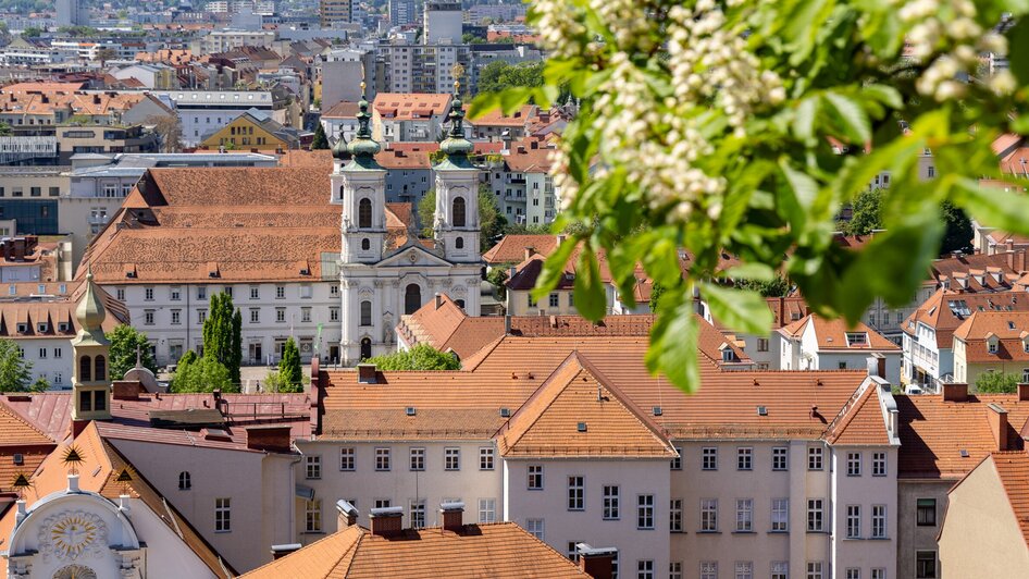 Blick über Graz mit der roten Dachlandschaft und der Mariahilferkirche im Hintergrund.