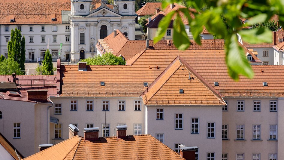 Blick über Graz mit der roten Dachlandschaft und der Mariahilferkirche im Hintergrund.