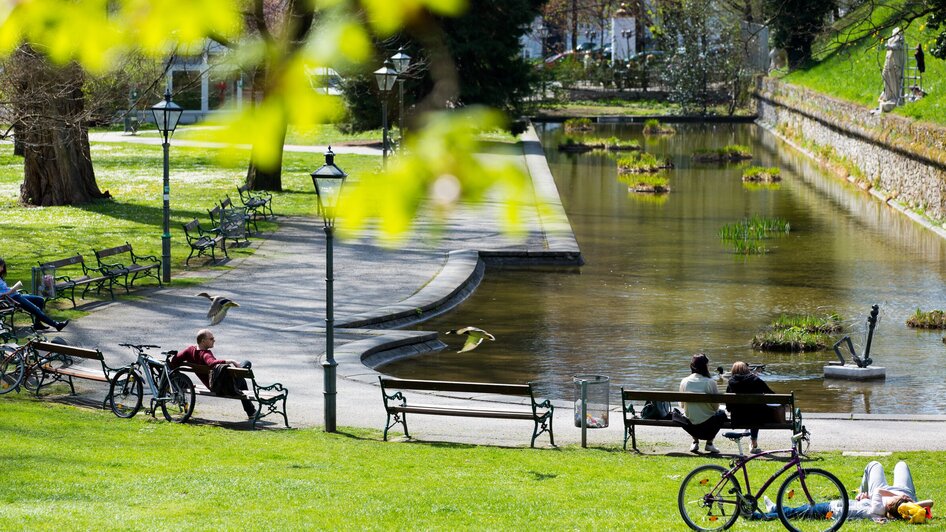 Ein ruhiger Stadtpark in Graz mit Bänken und einem Gewässer.