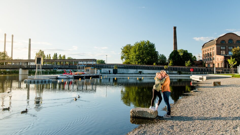 Mutter und Tochter umarmen sich am Ufer der Mur beim Stadtstrand in Graz bei Sonnenuntergang.