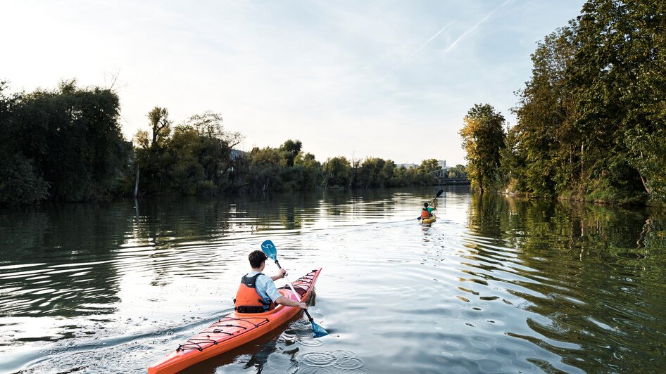 Zwei Personen im Kajak paddeln auf der ruhigen Mur umgeben von Bäumen. | © Stadtstrand Graz - Stefan Leitner