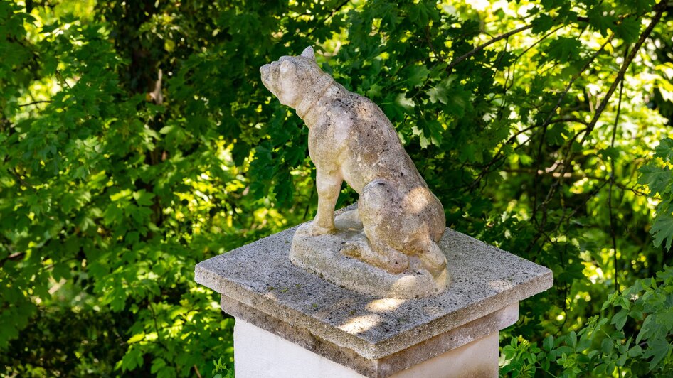 Eine Steinskulptur eines Hundes auf einem Podest, umgeben von grünen Blättern.