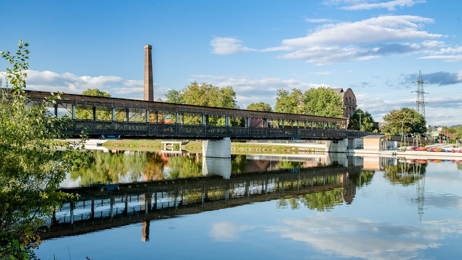 Eine Brücke über die Mur, idyllische Landschaft mit reflektierendem Wasser. | © Graz Tourismus - Mias Photoart