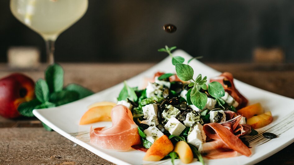 Frischer Salat mit Dressing, einem Getränk und einer Hand, die Zutaten hinzufügt. | © Julian Wallner