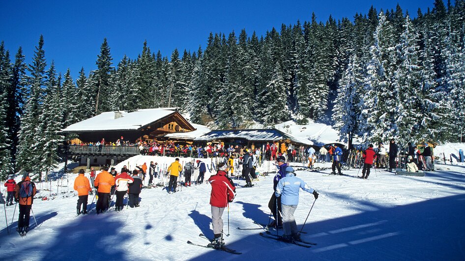 Seitenansicht einer Holzhütte umgeben von Skifahrern an der Piste | © Weitmoosalm