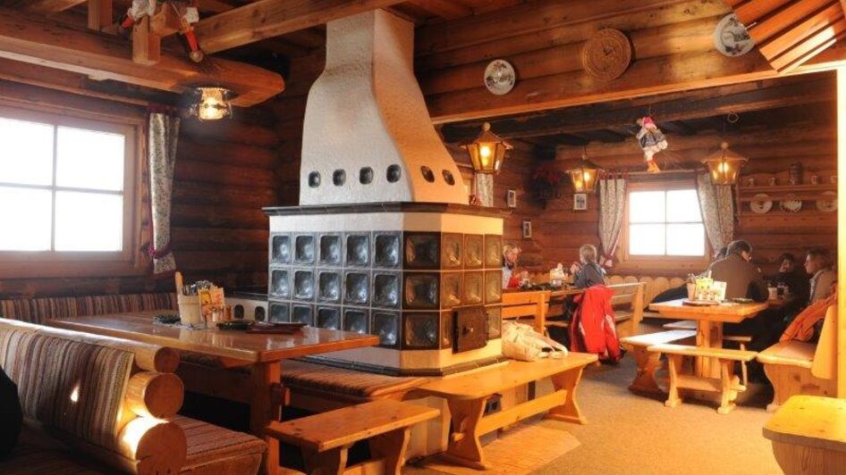Gemütlicher Gastraum mit Holzwänden, Polsterbänken, Holztischen und Kachelofen, warm beleuchtet. | © Weitmoosalm
